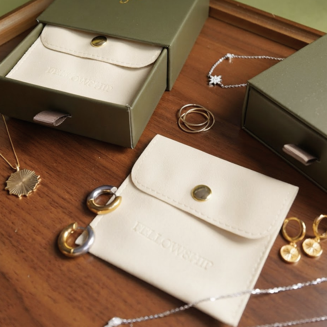 A styled flat-lay of jewelry accessories on a wooden surface, featuring a cream-colored leather pouch embossed with 'FELLOWSHIP', olive green jewelry boxes, and several gold and silver hoop necklaces and rings.