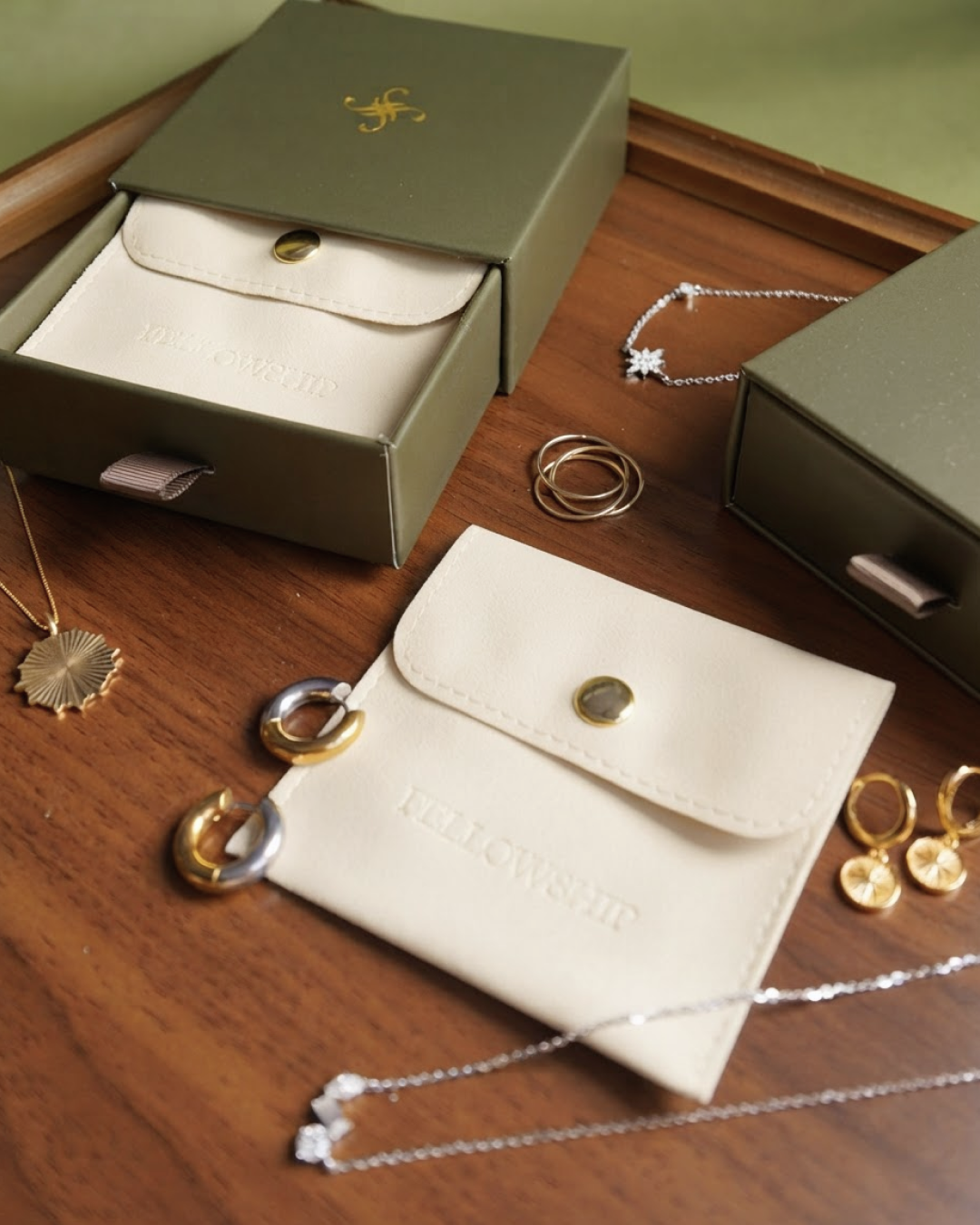 A styled flat-lay of jewelry accessories on a wooden surface, featuring a cream-colored leather pouch embossed with 'FELLOWSHIP', olive green jewelry boxes, and several gold and silver hoop necklaces and rings.