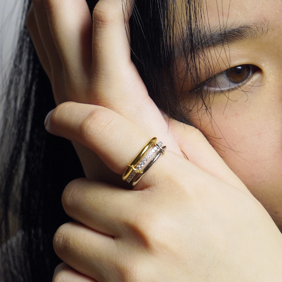 A close-up lifestyle shot of a person wearing a double-band ring on their finger. The ring features a polished gold band on one side and a silver band on the other, both adorned with small clear pavé-set stones.
